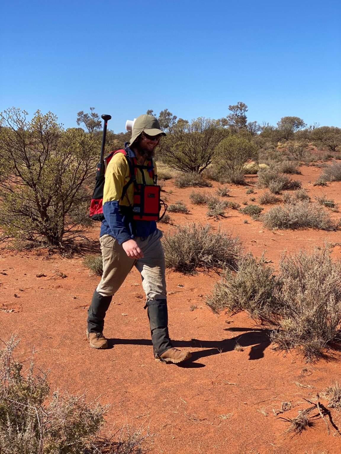 Operator in hi-vis walking through red-earth mulga scrub with the compact Modern Mag magnetometer backpack, sensor below head height